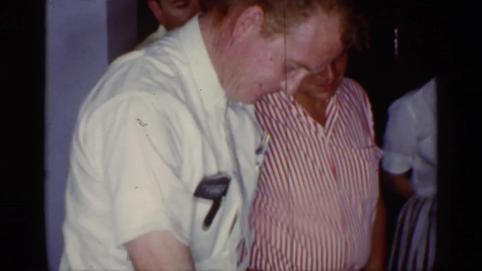 An older man in a white shirt stands beside a person in a red-and-white striped shirt during the indoor birthday gathering.