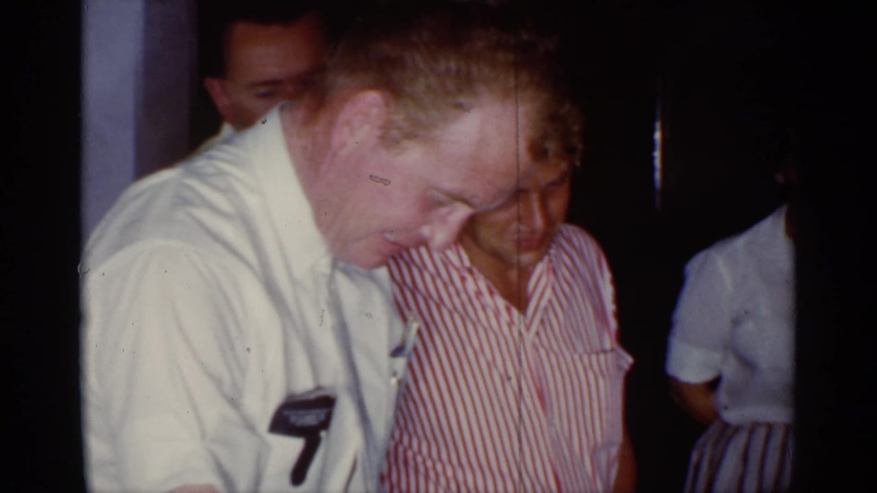 In 1959 Florida home-movie footage, adults in mid-century clothing gather indoors as a man in a light shirt looks down toward something off frame.