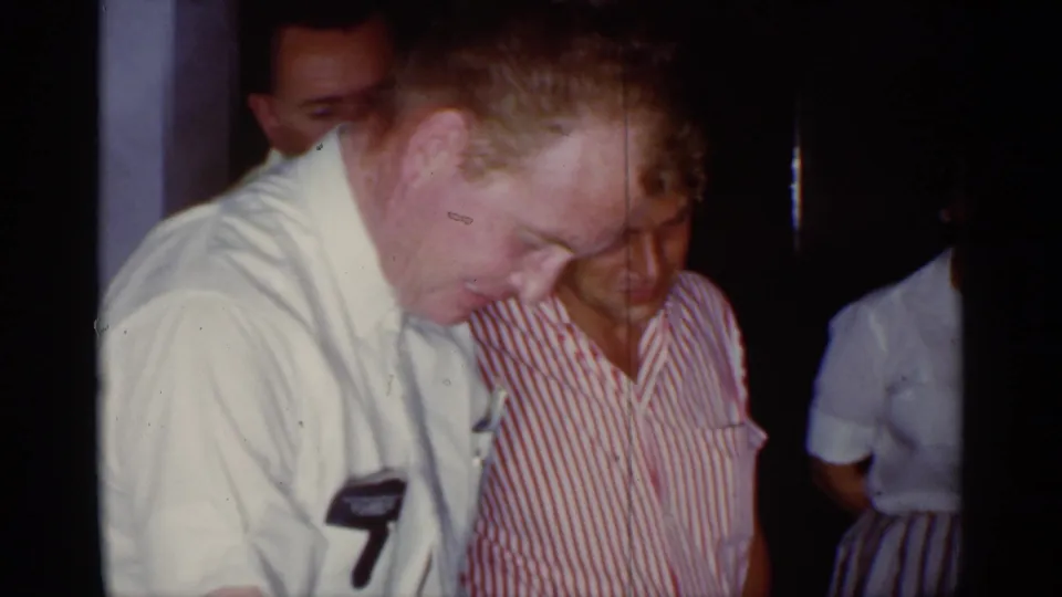 In 1959 Florida home-movie footage, adults in mid-century clothing gather indoors as a man in a light shirt looks down toward something off frame.