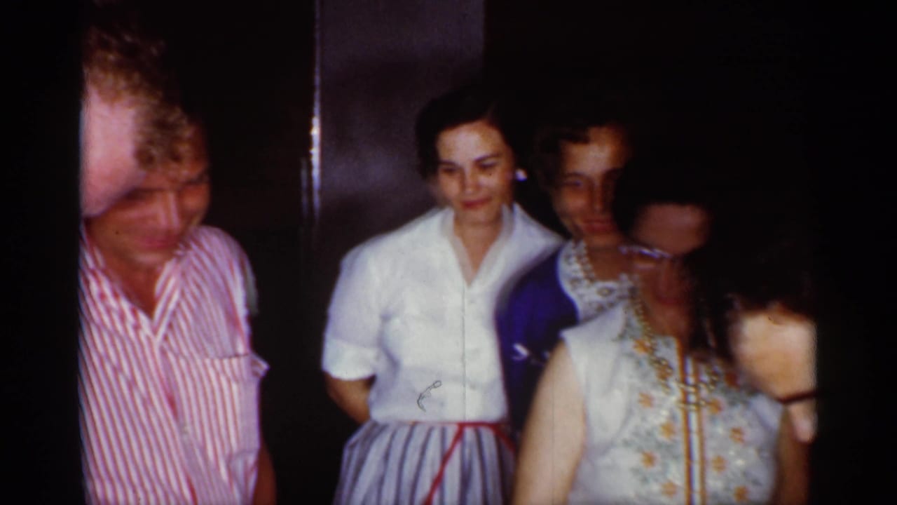 Women in dresses and blouses stand in a dim indoor space, watching toward the foreground.