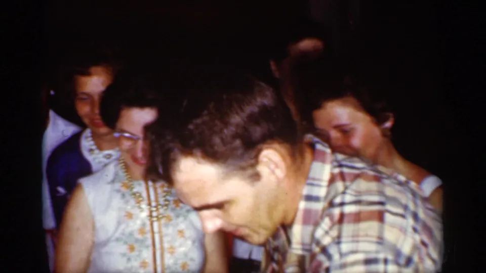 A man in a plaid shirt leans over something out of view while nearby adults watch in dim 1959 Florida home-movie footage.