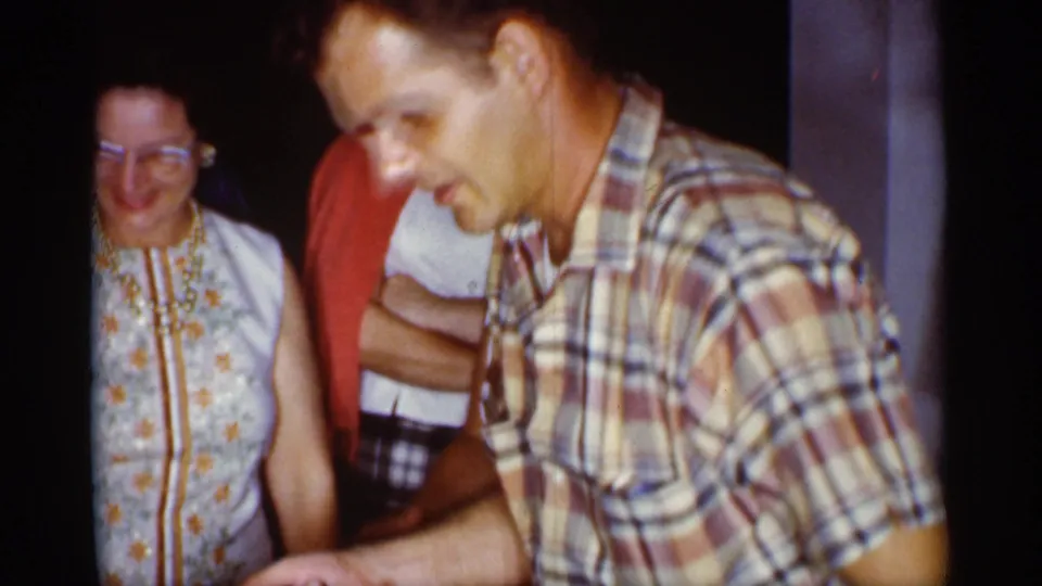 In a dim 1959 Florida interior, a man in a plaid shirt appears to present or handle an object while adults watch behind him.