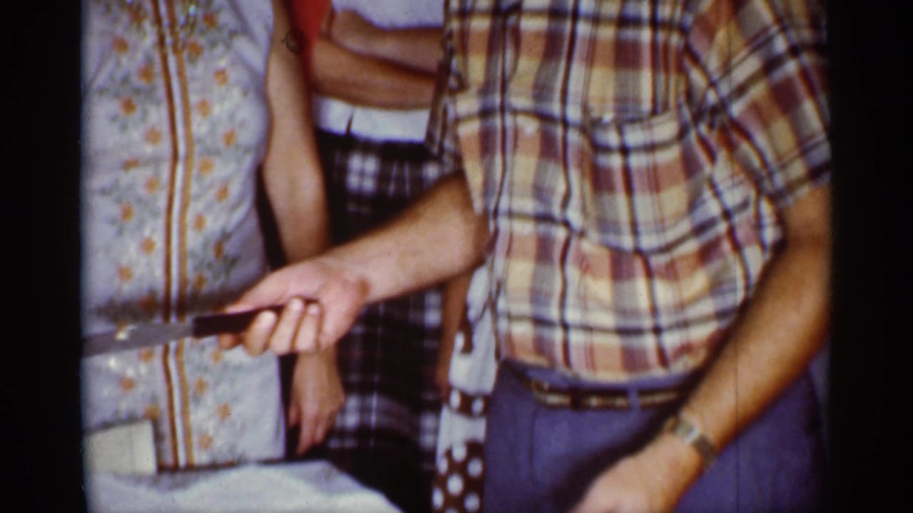 1959 Florida home-movie frame showing adults around a table while a man in a plaid shirt holds a knife before cutting cake.