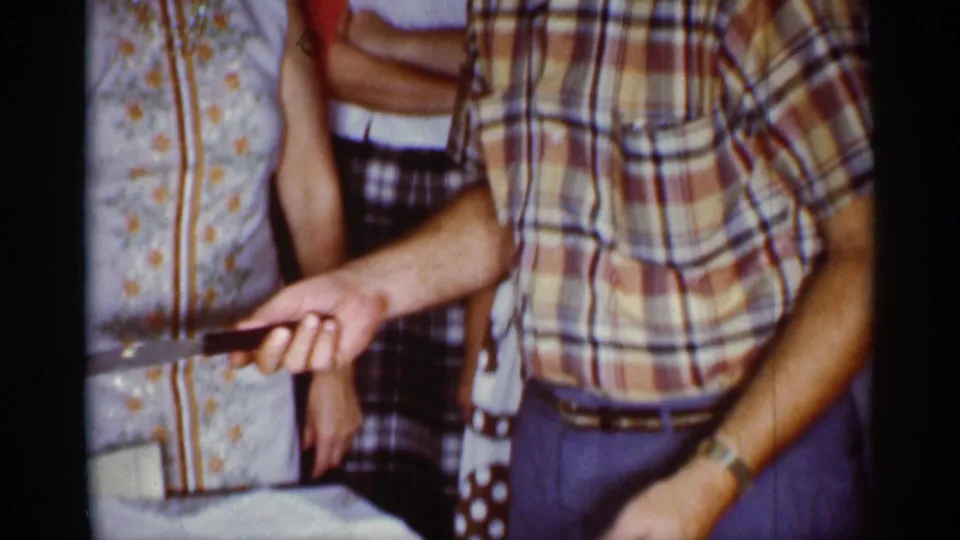 1959 Florida home-movie frame showing adults around a table while a man in a plaid shirt holds a knife before cutting cake.
