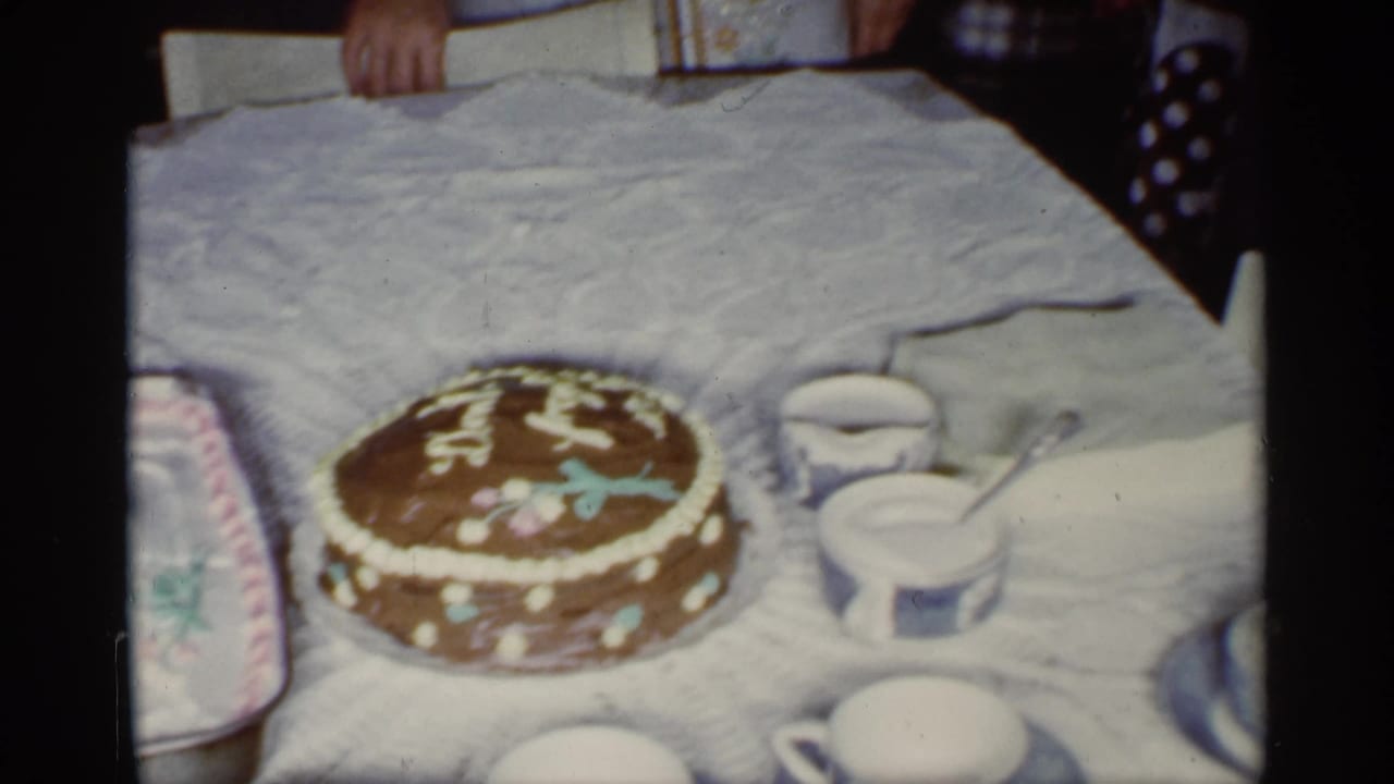 1959 Florida home-movie frame of a decorated round birthday cake on a white-covered table with cups nearby.