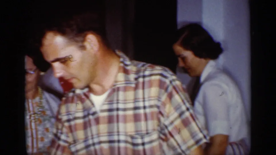 1959 Florida home-movie frame of adults gathered indoors, with a man in a plaid shirt foregrounded near the birthday table.