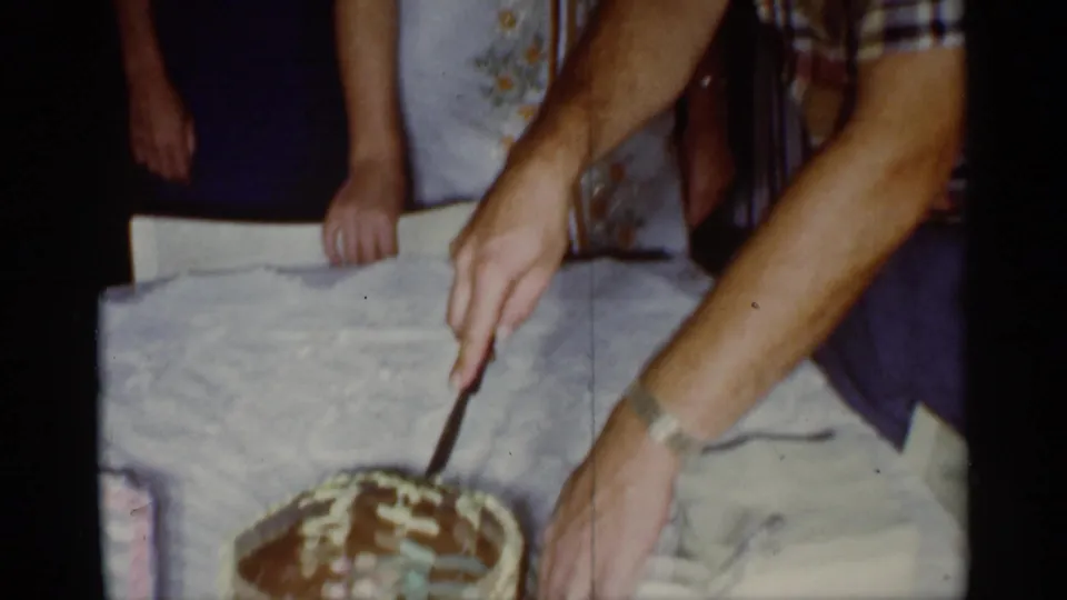 In a 1959 Florida home-movie frame, hands cut a chocolate birthday cake with white icing while relatives stand close around the table.