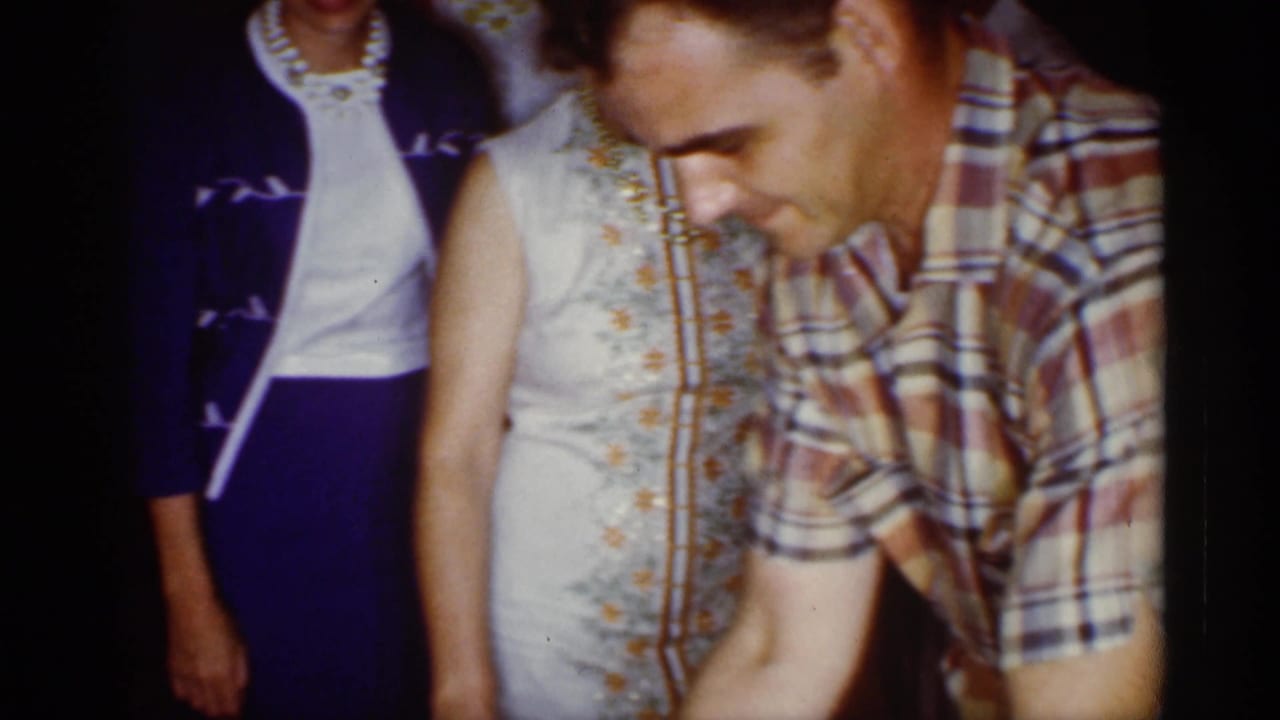 A man in a plaid short-sleeve shirt leans over the party table beside women in mid-century dresses during the Florida birthday gathering.