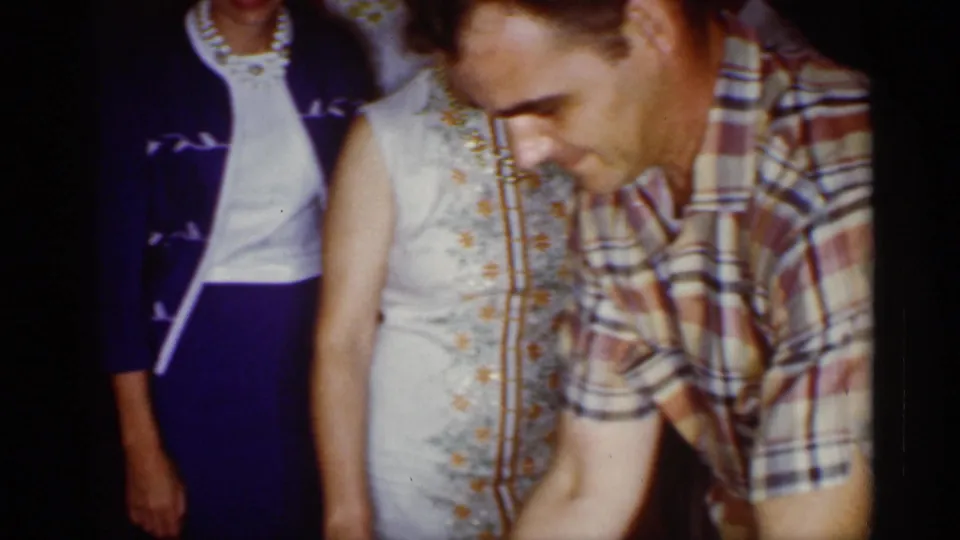 A man in a plaid short-sleeve shirt leans over the party table beside women in mid-century dresses during the Florida birthday gathering.