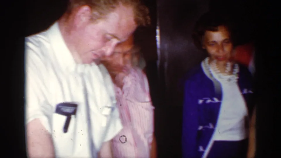 Family members stand in a dim private interior, with a man in a white shirt and a woman in a blue cardigan visible during the 1959 Florida birthday scene.