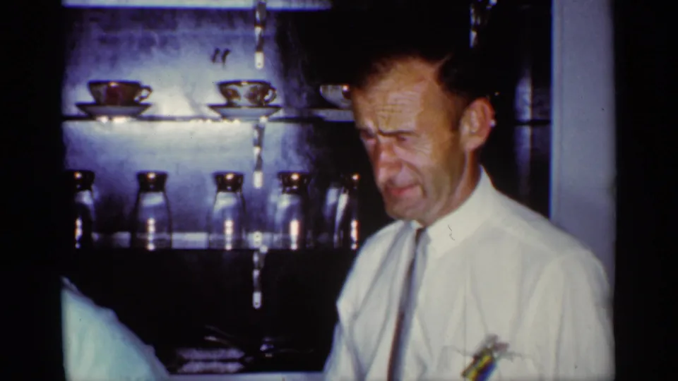 A man in a white shirt and tie stands in a kitchen-like interior with cups and glassware on shelves during the Florida home-movie birthday footage.