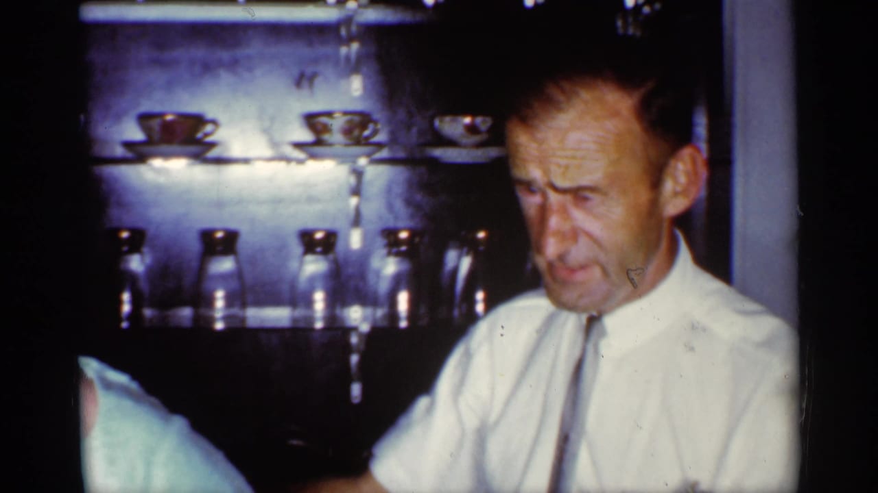 Man in white shirt and tie stands in a dim kitchen near shelves of cups and jars.