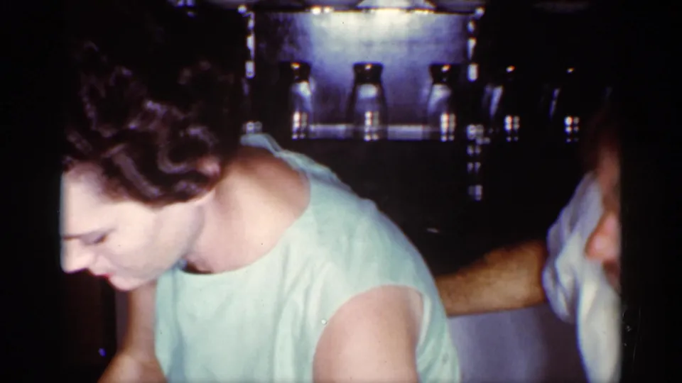 1959 Florida home-movie frame showing a woman in a pale green dress leaning over a kitchen table as a man stands partly in view.