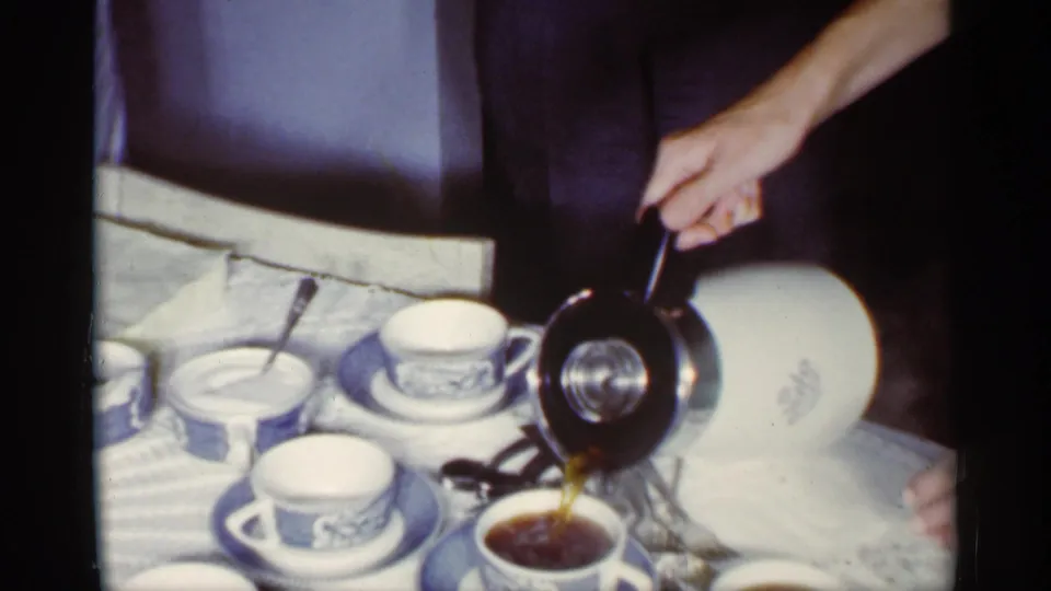 1959 Florida home-movie frame showing coffee poured from a white pot into blue and white cups with saucers on a kitchen table.