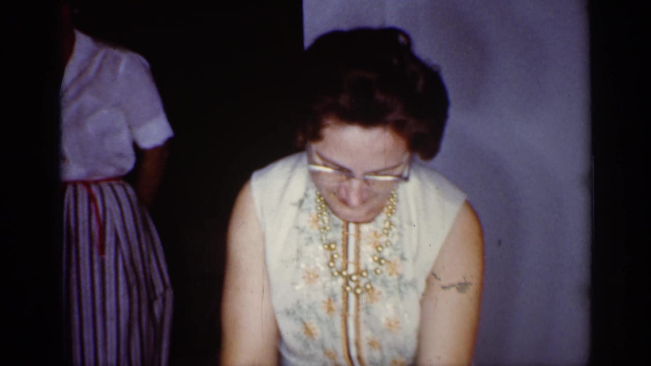 Woman in glasses and a sleeveless floral dress looks down indoors.