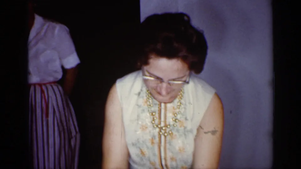 Woman in glasses and a sleeveless floral dress looks down during a 1959 Florida home gathering, with another guest partly visible behind her.