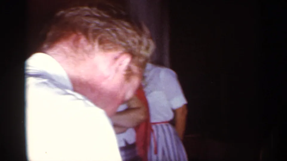 A man leans forward in a dim 1959 Florida home-movie frame, with other guests partly visible around him.