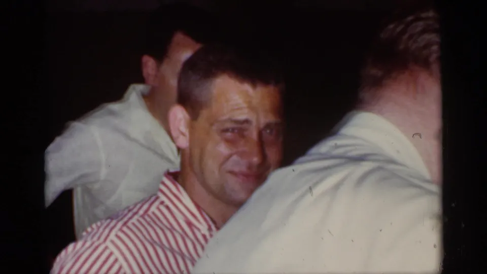 Man in a red and white striped shirt sits among guests in a dim private interior in 1959 Florida home-movie footage.
