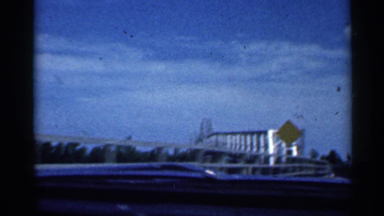 1959 Indiana 8mm frame from inside a moving car approaching a pale metal truss bridge under a blue sky.