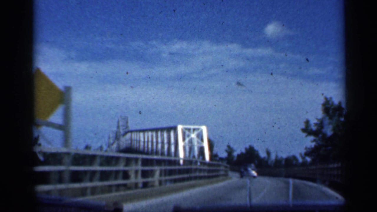 Car windshield view on a bridge roadway with white trusswork and roadside railings.