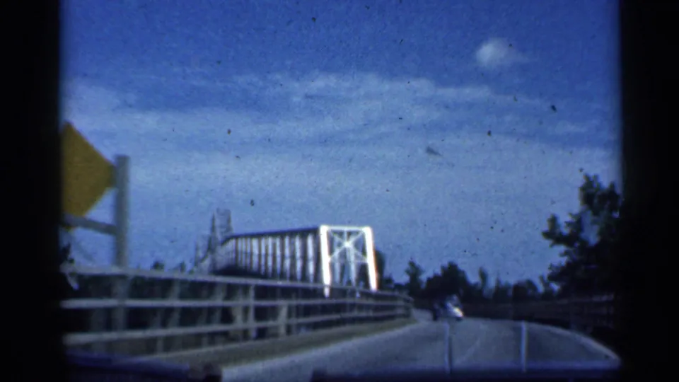 Inside-car view crossing a white metal bridge in Indiana, with railings, trusswork, trees, and sky visible.
