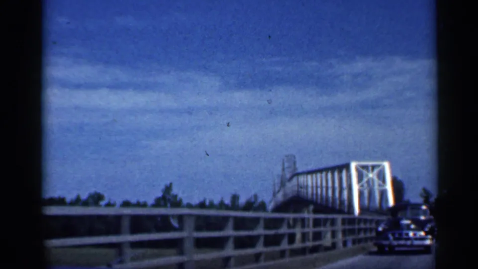 1959 Indiana road view from inside a car, showing bridge railings, a white truss span, and a vehicle near the right edge.