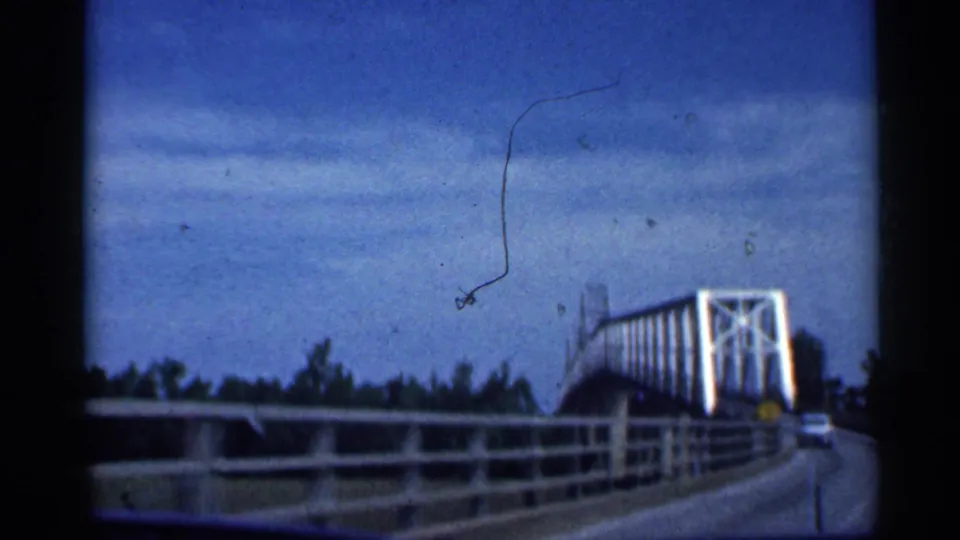 Inside-car view near the end of the Indiana bridge crossing, with trusswork, railings, trees, and a passing vehicle visible.