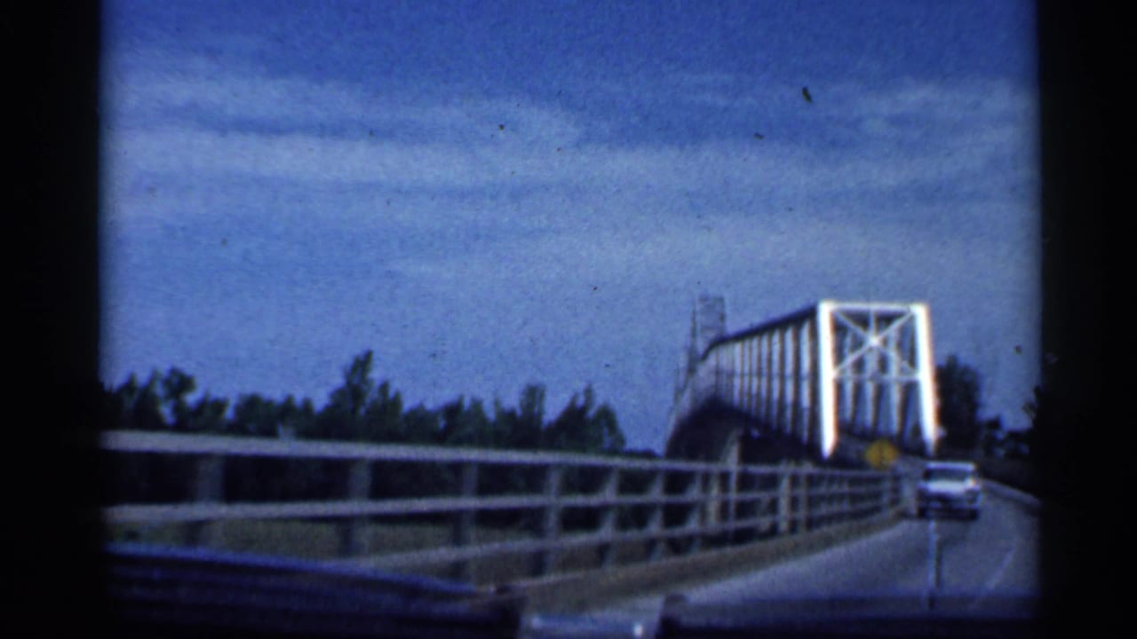 Vehicle approaches a white steel truss bridge as an oncoming car nears the narrow roadway.