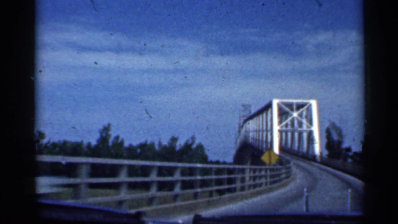 1959 Indiana home-movie frame showing a vehicle approach to a white steel truss bridge, with guardrails and a yellow warning sign beside the road.