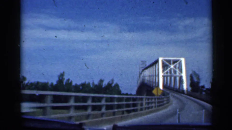 1959 Indiana home-movie frame showing a vehicle approach to a white steel truss bridge, with guardrails and a yellow warning sign beside the road.