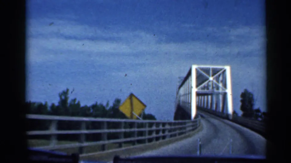 1959 Indiana 8mm frame from inside a moving vehicle nearing the white steel truss bridge, with a yellow sign and roadside guardrail visible.