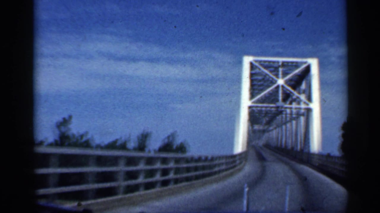 Car approaches a white steel truss bridge on a sunny road.