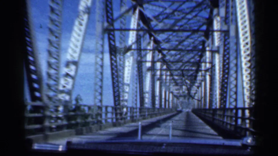 Inside the bridge span, the camera looks forward through repeating steel trusses and narrow roadway lines.