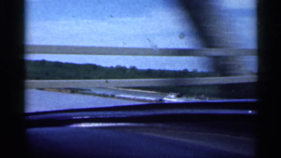 From the moving car, the camera catches the river or waterway and tree-lined shore through bridge railings.