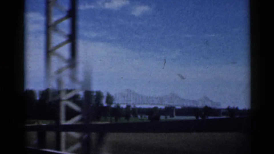 A moving 8mm view shows a roadside structure, low riverbank, and distant bridge under a bright Indiana sky.