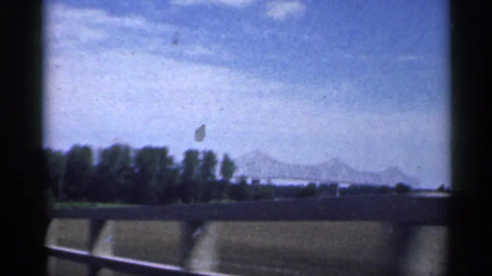 The final 1959 Indiana frame shows trees, a distant bridge, road barrier, and pale sky from a passing vehicle window.