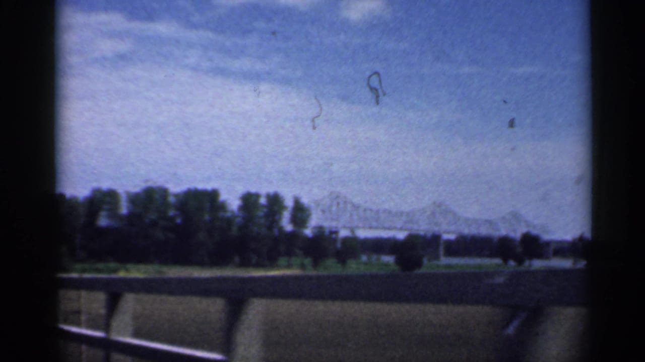 Car-window view of a highway barrier, trees, sky, and a distant truss bridge.