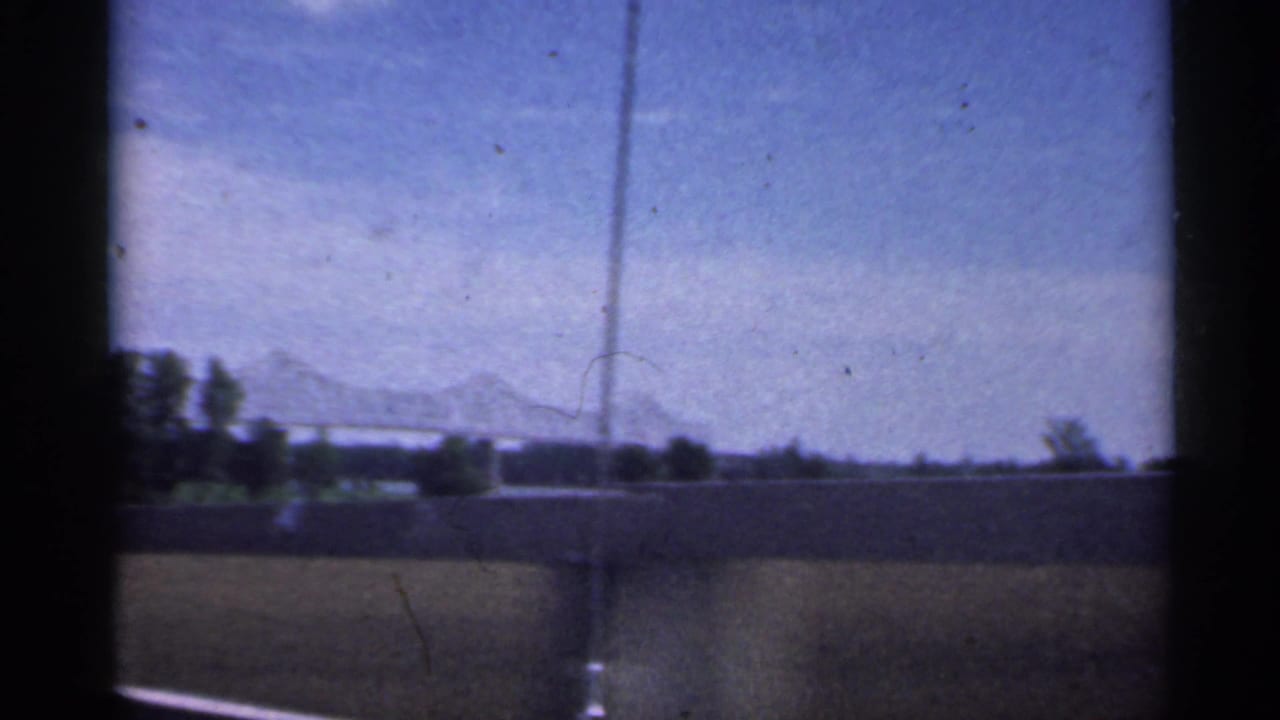 1959 Indiana home-movie frame with a road or bridge edge in the foreground and a distant truss bridge across the horizon.