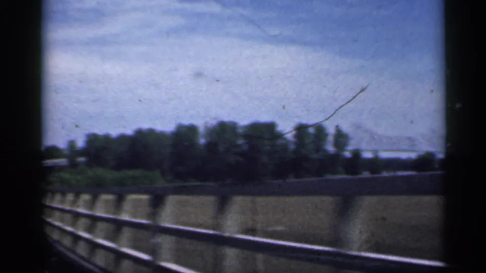 1959 Indiana 8mm side view from a moving vehicle, with guardrail, concrete barrier, trees, and a faint bridge line beyond.