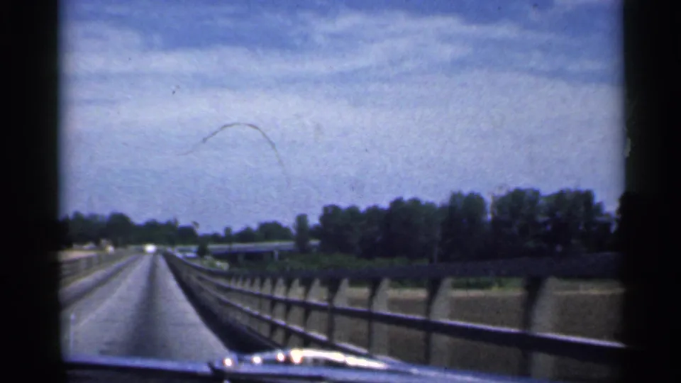 1959 Indiana 8mm frame looking forward along a highway bridge, with guardrails, concrete sides, trees, and open sky ahead.