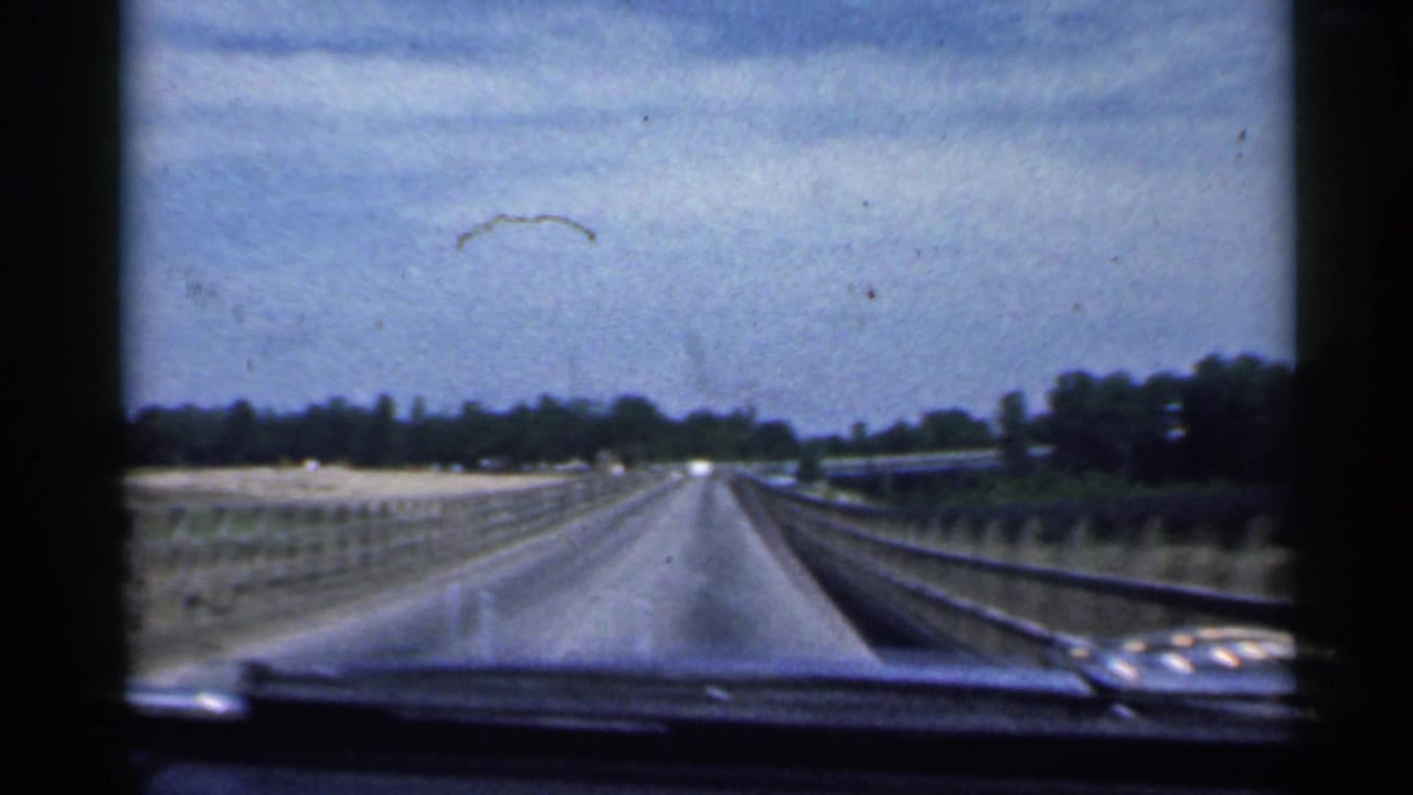 1959 Indiana home-movie view from inside a car approaching a long bridge, with road, rails, dashboard edge, and trees visible.