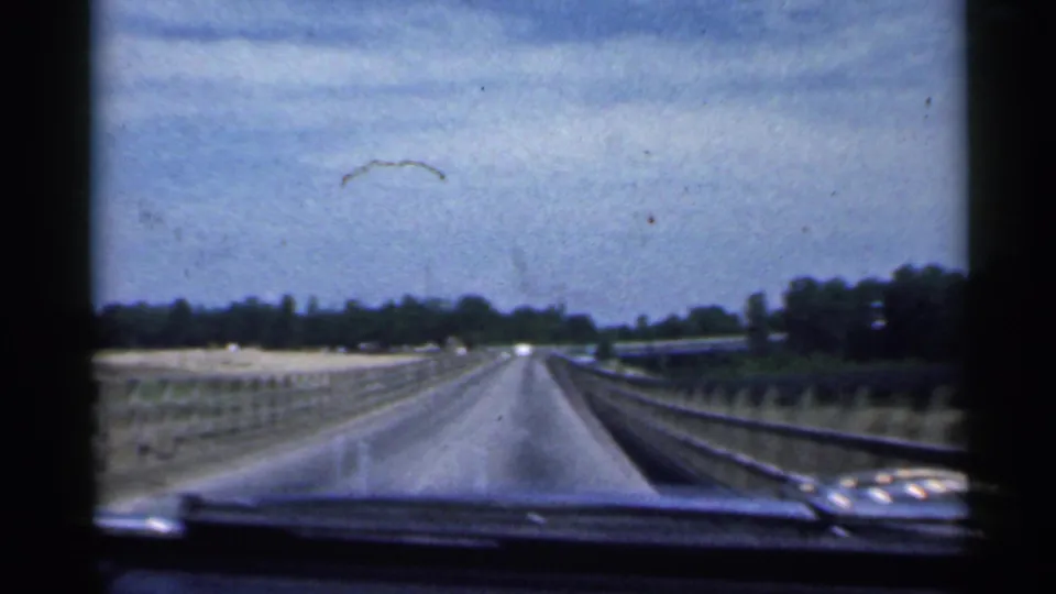 1959 Indiana home-movie view from inside a car approaching a long bridge, with road, rails, dashboard edge, and trees visible.