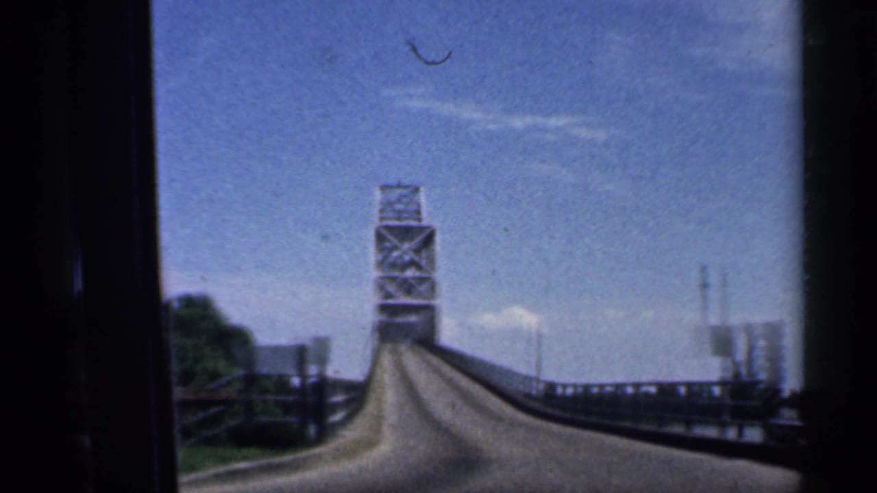 Steel truss bridge ahead on a raised roadway beside water and railings.