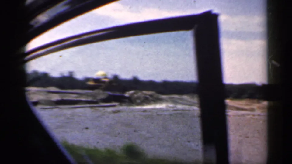 From inside the car, a fishing rod crosses the foreground while water, shoreline, and distant trees pass outside.