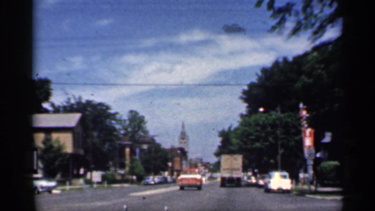 1959 Indiana 8mm frame from inside a car, looking past traffic toward a church clock tower on a tree-lined street.