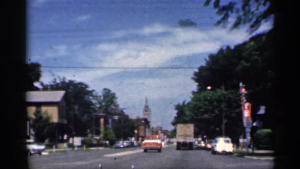1959 Indiana 8mm frame from inside a car, looking past traffic toward a church clock tower on a tree-lined street.