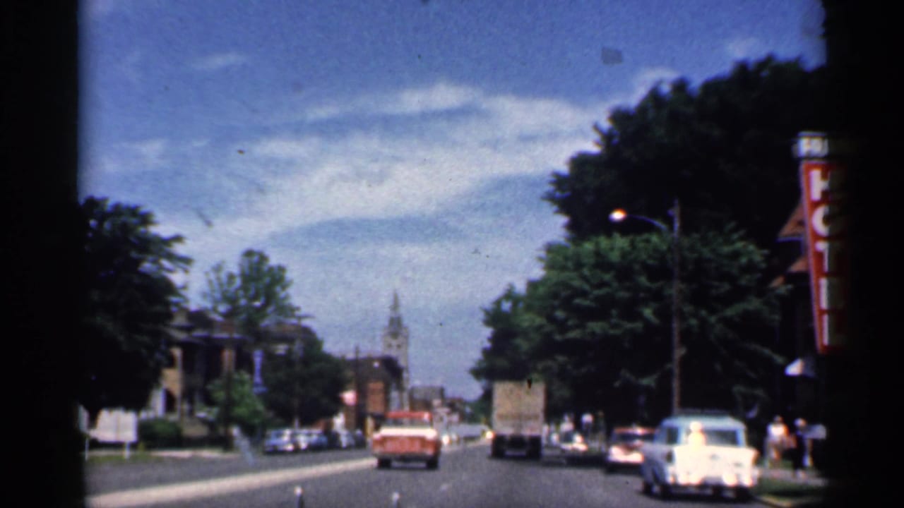 Moving 8mm view in Indiana, 1959, with mid-century cars, a hotel sign, trees, and a distant church tower.