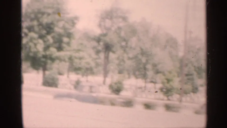 1959 Indiana 8mm frame looking through a dark car-window divider toward a washed-out tree-lined street or roadside.