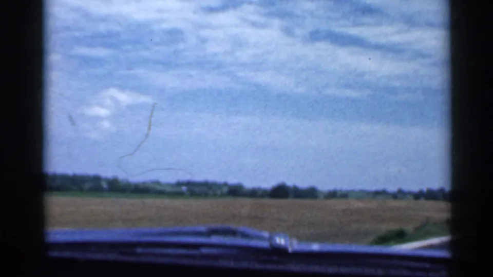 1959 Indiana countryside view from inside a car, with dashboard foreground, open field, distant tree line, and blue sky.