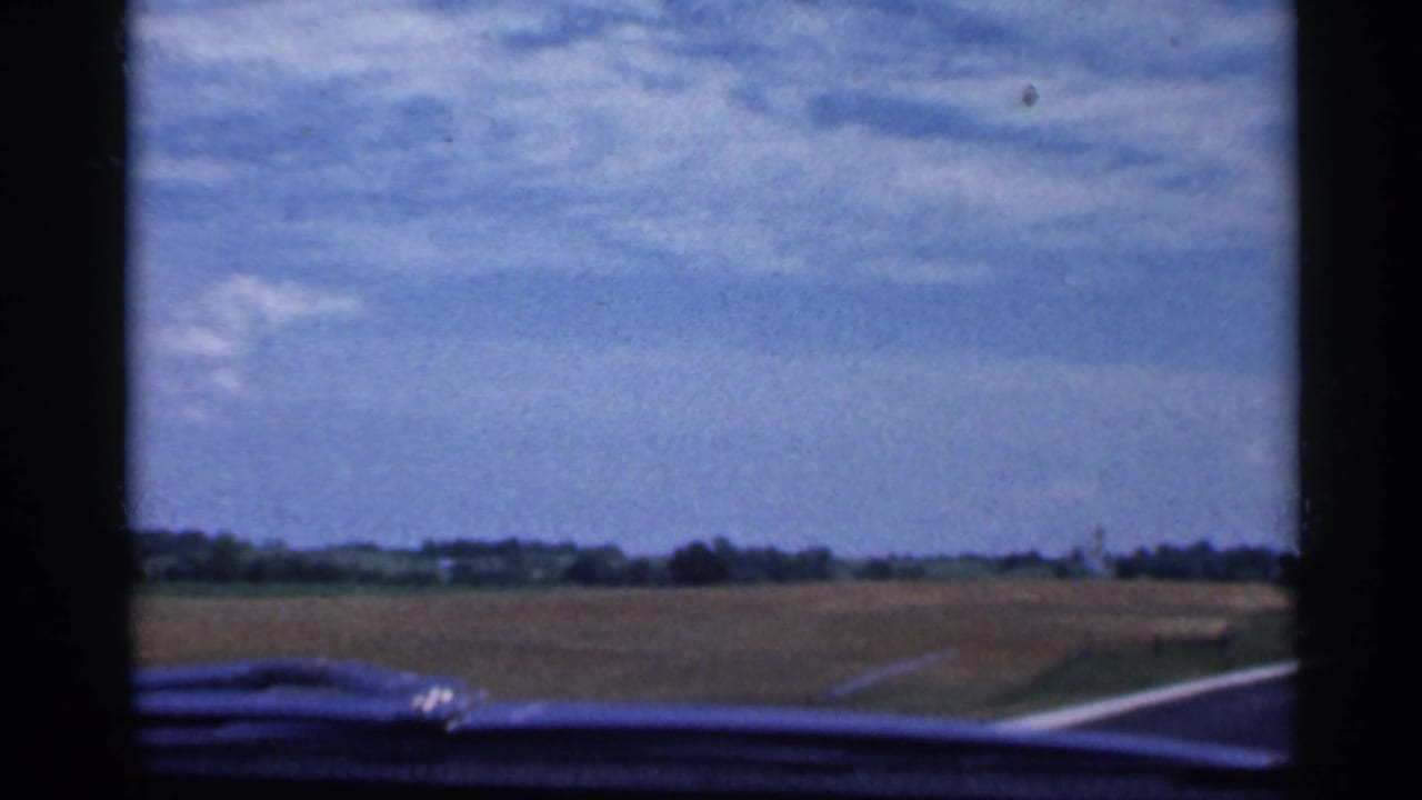 Rural road seen through a car windshield with fields and a blue sky.